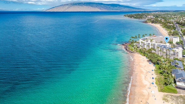 Luftaufnahme einer Küstenregion mit Strand, klarem blauen Wasser und einem Berg im Hintergrund.