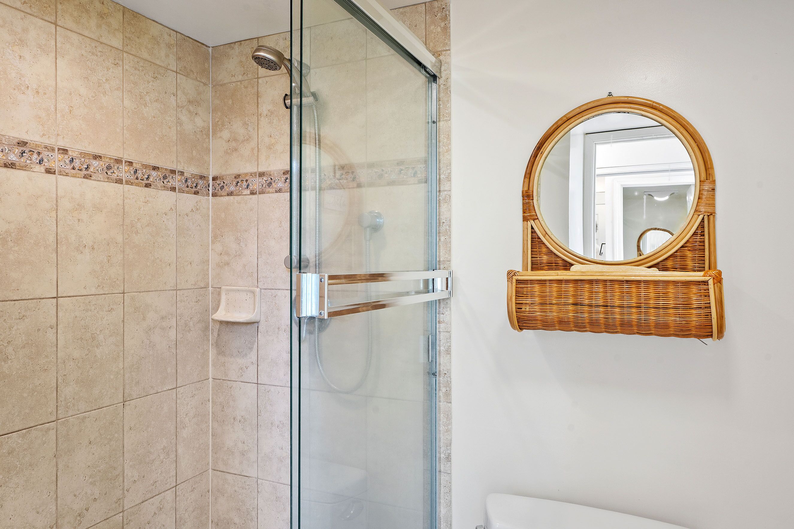 Un baño con ducha de vidrio, inodoro blanco y estante de mimbre con espejo redondo.