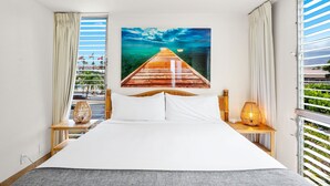 Ein Hotelzimmer mit einem großen Bett, einem Gemälde eines hölzernen Piers, Meerblick und einem Balkon mit Vorhängen.