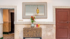 Une pièce dotée d’un sol carrelé, d’une porte en bois, d’un miroir avec un cadre décoratif, d’une petite table sur laquelle est posé un vase de fleurs et d’un tapis à motifs.