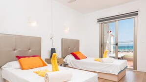 A hotel room with two beds, each with a white bedspread and orange pillows, a small table with a lamp, and a view of the beach through a sliding glass door.