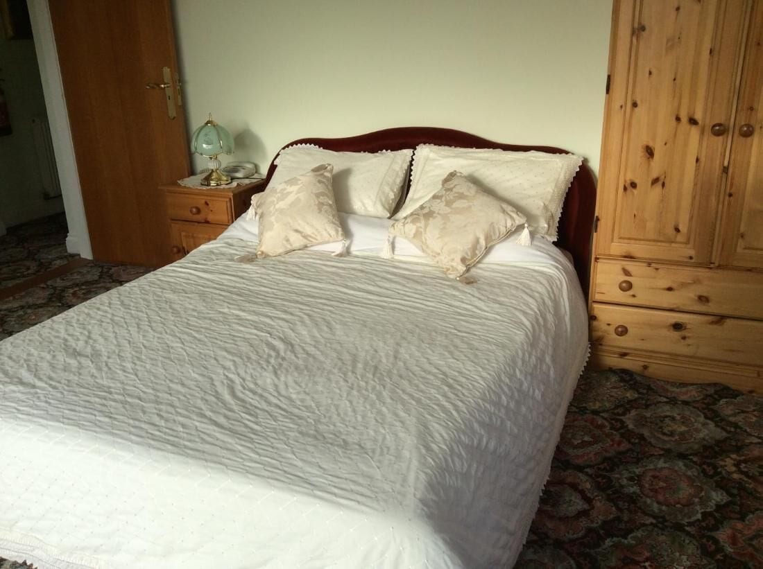 Ein ordentlich gemachtes Bett mit weißer Decke und vier beigen Kissen, ein Nachttisch aus Holz mit einer Lampe sowie ein Holzschrank mit mehreren Schubladen.