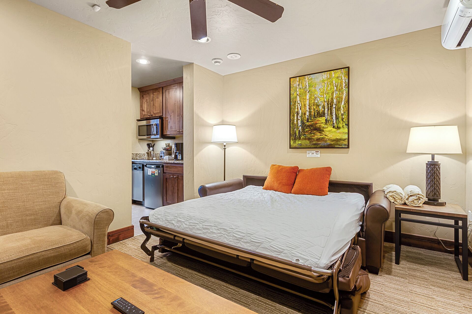 A hotel room with a bed, a sofa, a desk, a lamp, a painting, and a kitchenette.