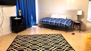 Quarto com cama, mesa de cabeceira, abajur, cortina azul e tapete estampado.
