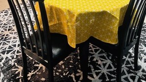 A dining area with a yellow tablecloth, wooden chairs, a vase with dried flowers, and a microwave on the counter.