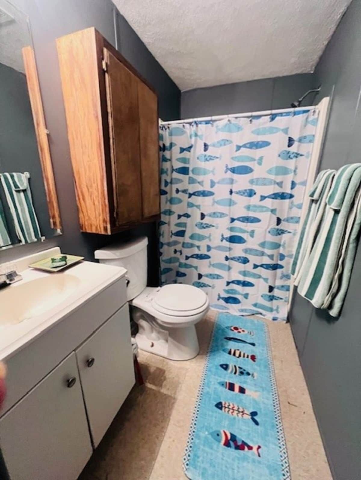 Une salle de bain comprenant des toilettes, un lavabo, un meuble en bois, un rideau de douche orné de motifs de poissons et un tapis bleu à motifs de poissons.