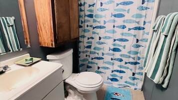 Une salle de bain comprenant des toilettes, un lavabo, un meuble en bois, un rideau de douche orné de motifs de poissons et un tapis bleu à motifs de poissons.
