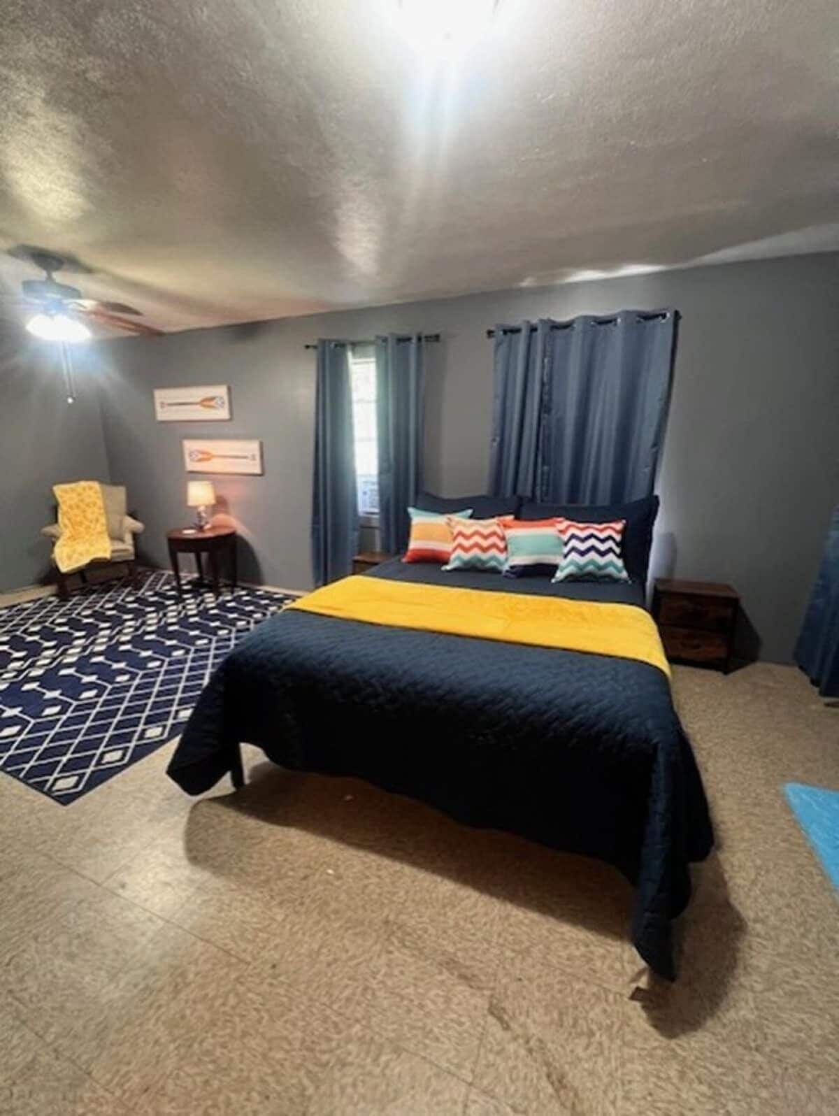 A bedroom with a bed, curtains, a chair, a lamp, and a rug.