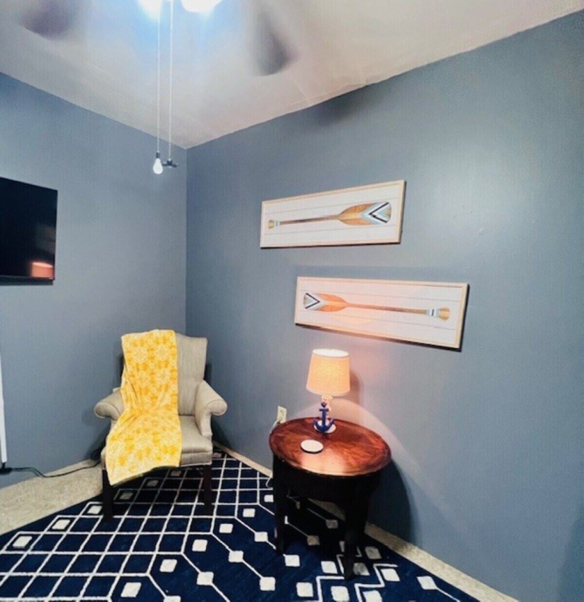 Una habitación con piso estampado, una silla con una manta amarilla, una pequeña mesa redonda con una lámpara y dos cuadros colgados en la pared.