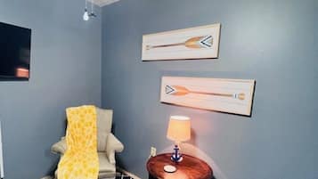 Una habitación con piso estampado, una silla con una manta amarilla, una pequeña mesa redonda con una lámpara y dos cuadros colgados en la pared.