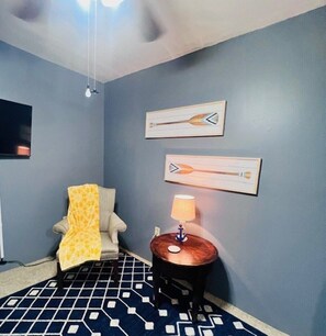 A room with a patterned floor, a chair with a yellow blanket, a small round table with a lamp, and two wall-mounted paintings. - Rachel's Place (Vicksburg)