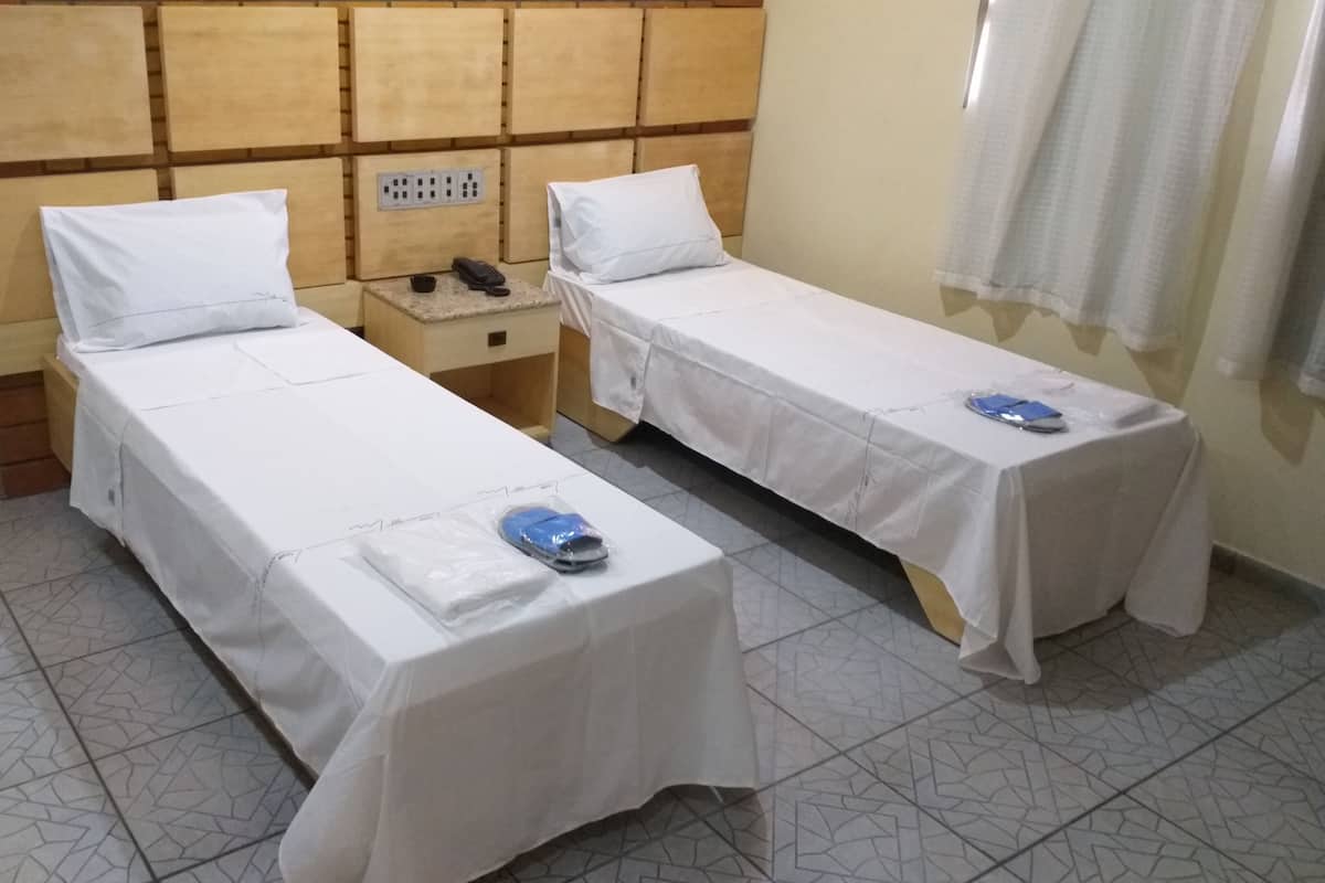 Two single beds with white linens, a wooden headboard, a bedside table with a phone and a cup, and a tiled floor.
