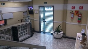 Una zona de recepción con suelo de mármol, un mostrador de recepción, un televisor, un extintor y una planta en maceta.