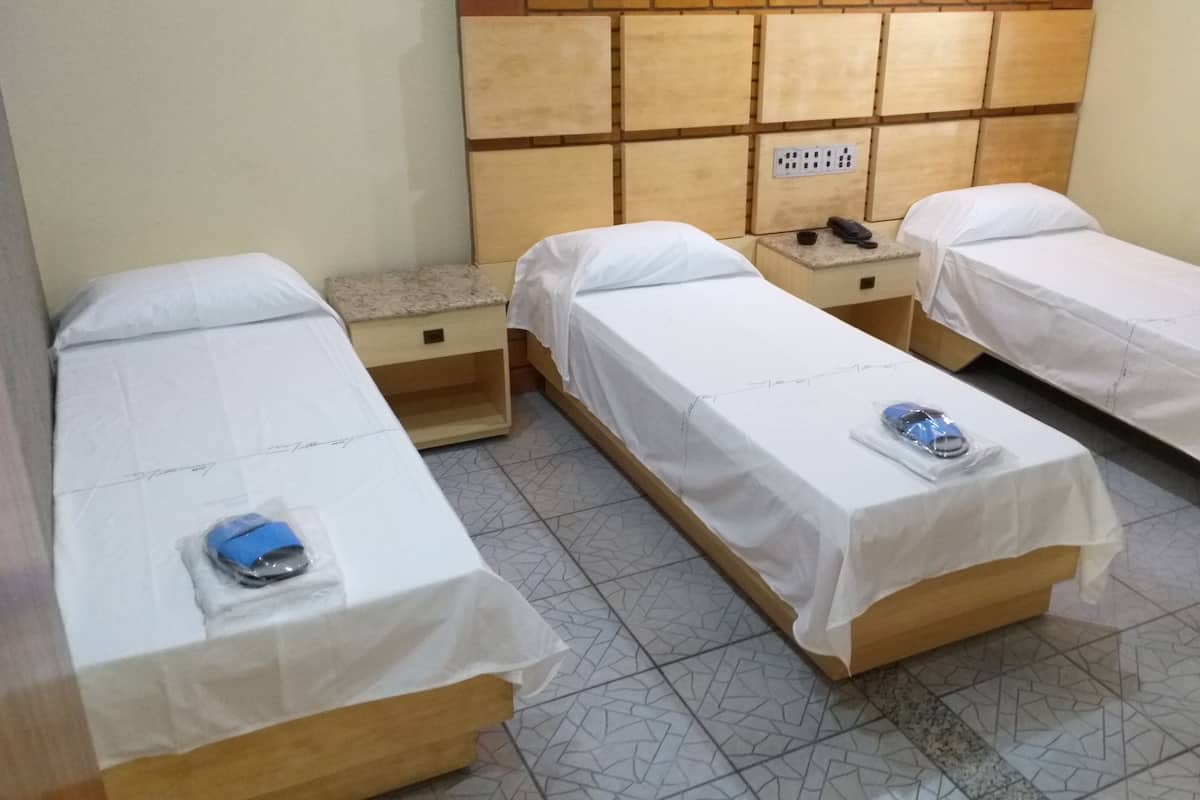 A hotel room with three single beds, each with a white bedsheet and a small item on the bed. There is a wooden headboard with a power outlet and a telephone on the nightstand.