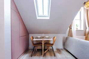 Een kleine, goed verlichte kamer met een schuine kap, een eettafel met stoelen, een bank en een raam met gordijnen.