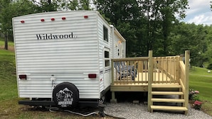 A white RV with 'Wildwood' branding parked on gravel, connected to a wooden deck with a ramp.