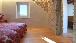 A bedroom with a wooden floor, a bed with a patterned bedspread, a window, and a stone wall. - Baita Tersilia Near Lake And Mountain  (Borgo Valbelluna)
