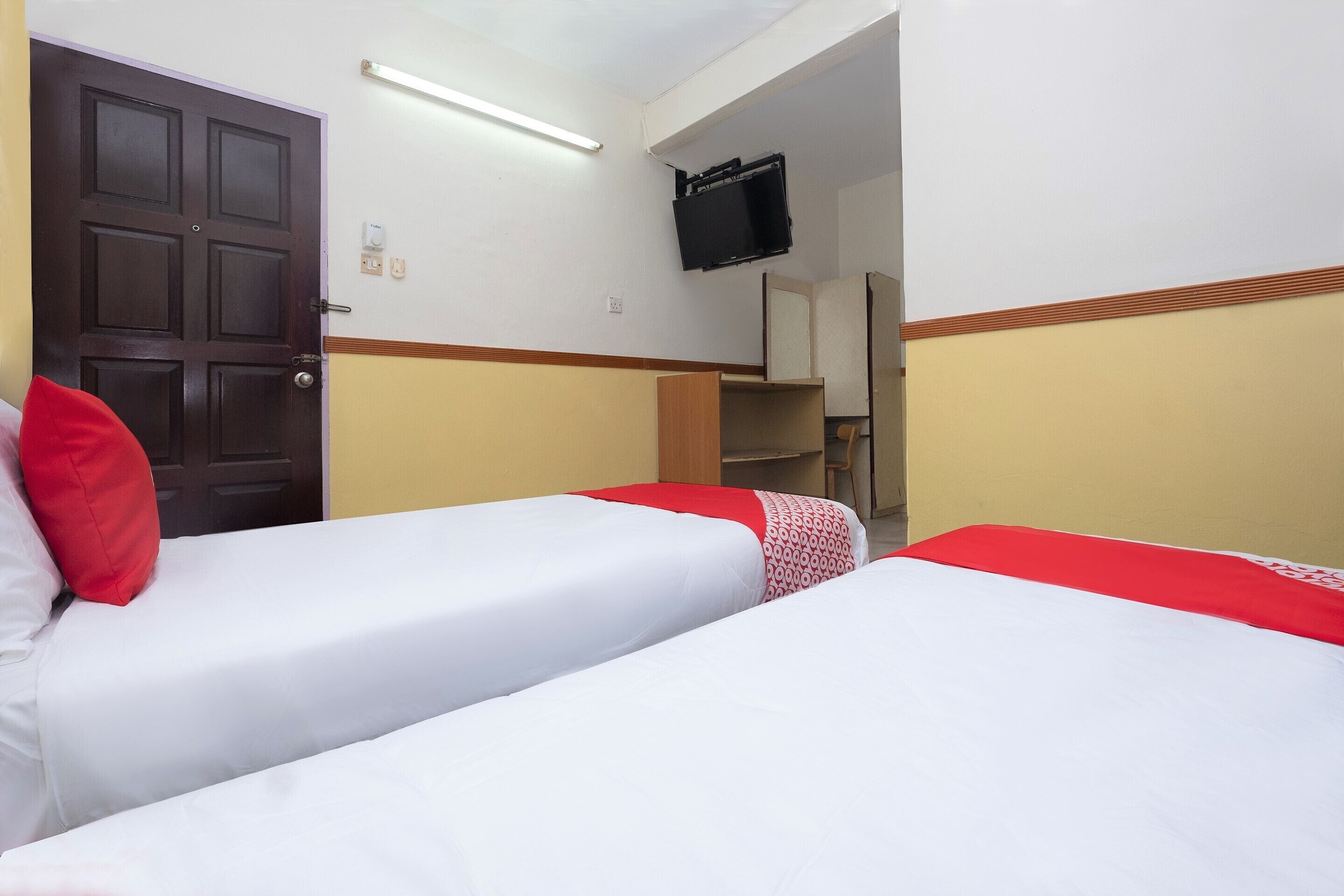 A hotel room with two beds, one with a red and white patterned bedspread and the other with a plain white bedspread. There is a wooden door, a television mounted on the wall, and a small shelf with items on it.