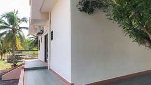 Ένα κτίριο με επίπεδη στέγη, μια πλακόστρωτη είσοδο και έναν μικρό κήπο με φοίνικες.