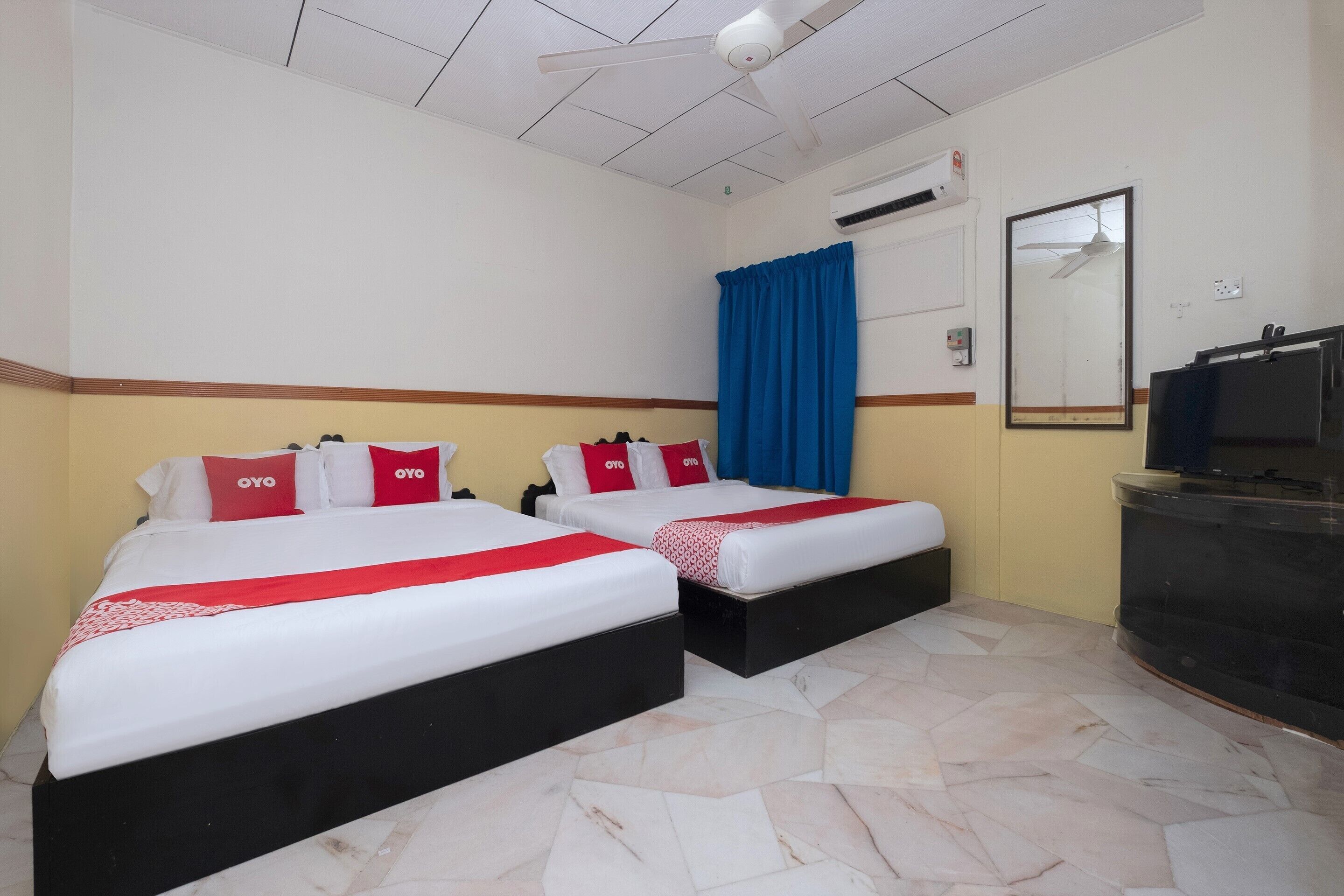 A hotel room with two beds, each with red and white bedding and pillows, a blue curtain, a flat-screen TV, and a tiled floor.