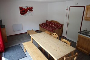 A dining area with a wooden table and benches, a red patterned sofa, a kitchen area with wooden cabinets, and a wall-mounted artwork of red flowers.