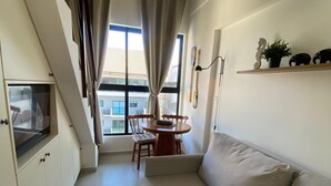 Ein modernes Wohnzimmer mit einer Couch, einem Esstisch mit Stühlen, einer Küchenzeile mit Schränken und einem großen Fenster mit Vorhängen.
