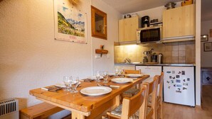 Een eethoek met een houten tafel voor vier personen, een keuken met houten kastjes en een magnetron, en een koelkast met magneetjes.
