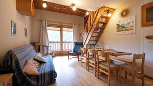 Een gezellige kamer met een houten trap, een eettafel met stoelen, een bank met kussens en een groot raam met gordijnen.