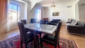 A spacious living room with a dining area, large windows, and a ceiling fan. - Bright air-conditioned flat in the city centre (Nice)