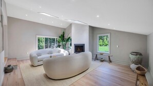 Ett modernt vardagsrum med en stor vit soffa, en öppen spis och stora fönster med utsikt över grönområden.