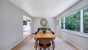 A dining room with a wooden table, chairs, a vase with flowers, and a large window with a view of trees.