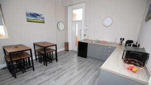 Een moderne keuken met grijze kastjes, een houten eettafel met stoelen, een koffieapparaat en een fruitmand op het aanrecht.