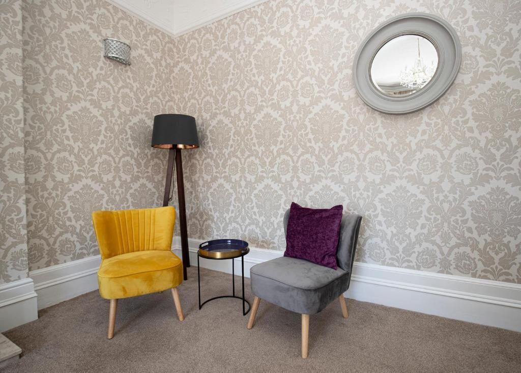A room with two chairs, one yellow and one grey, a small round table, a floor lamp, and a round mirror on the wall.