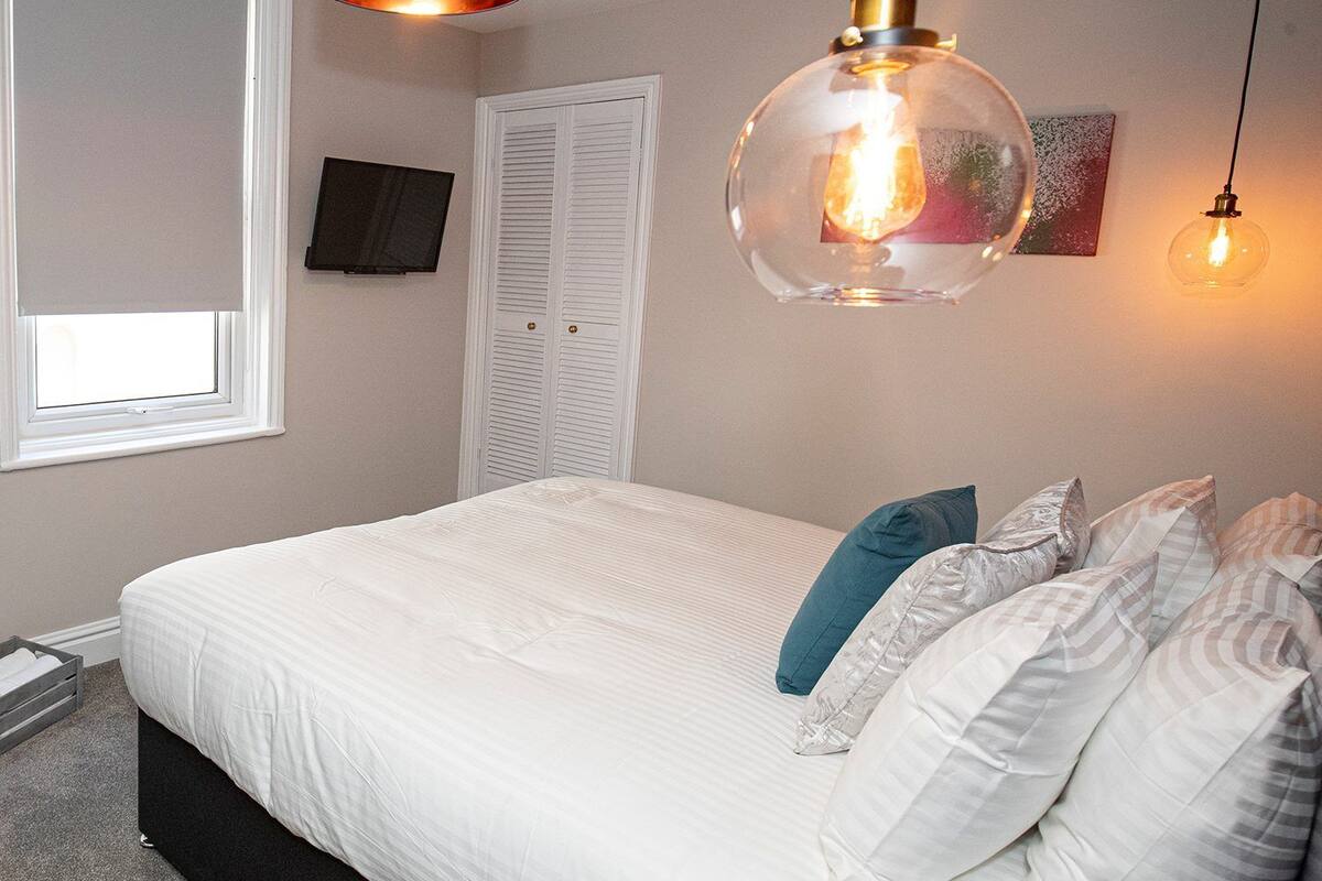 Un dormitorio con una cama, ventana, televisor, puerta y tres lámparas colgantes.