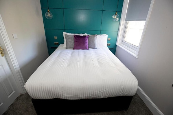 Una cama bien tendida con edredón y almohadas blancos, una pared de acento color aguamarina y una ventana que deja entrar luz natural.