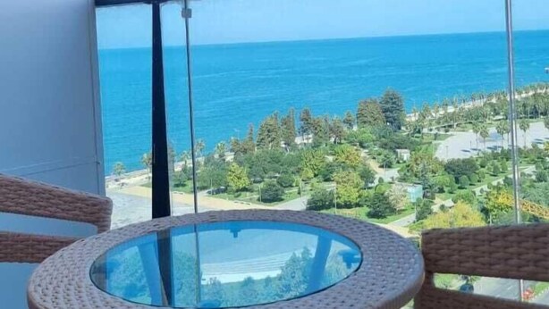 A balcony with wicker chairs and a glass table, overlooking a coastal view with the ocean and greenery. - Tal's Apartment  (Batumi)