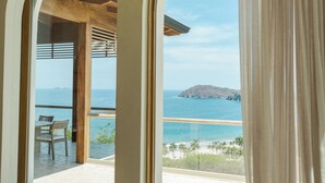 Una habitación con un amplio ventanal arqueado que da a una playa y al océano, con techo de madera y una mesa con sillas.