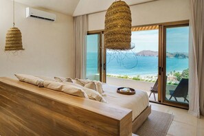 A bedroom with a wooden bed, a large window with a view of the sea, and a balcony with a chair.