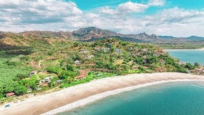 Un complejo turístico costero con varias construcciones, una playa y montañas al fondo.