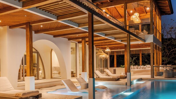 Una casa moderna de varios niveles con piscina, mobiliario de exterior e interiores bien iluminados.