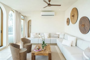 A modern living room with a sofa, coffee table, and chairs. There is a ceiling fan and an air conditioning unit.