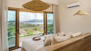 A bedroom with a large bed, a wooden bench, a woven light fixture, and a view of a landscape through a sliding door. - Ocaso Culturally Rich Villa, Flamingo Beach (Flamingo)