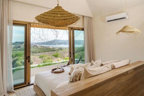 Une chambre avec un grand lit, un banc en bois, un luminaire en fibres tressées et une vue sur un paysage, visible à travers une porte coulissante.