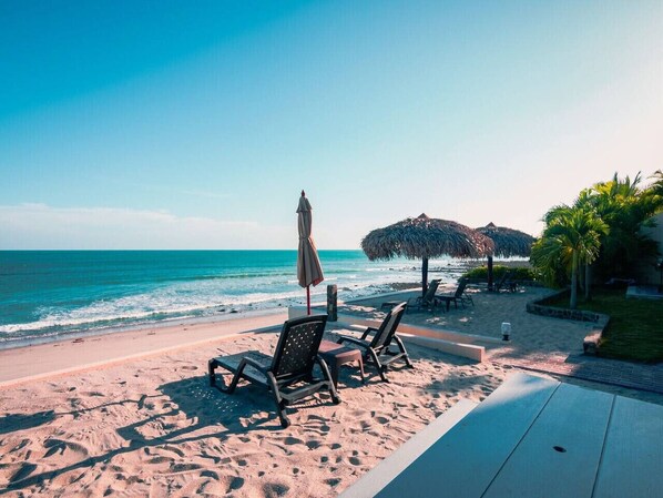 A beach with lounge chairs and umbrellas, a thatched structure, and palm trees.