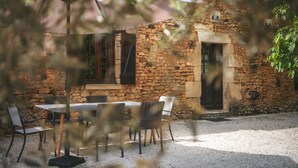 Ein Sitzbereich im Freien mit Tisch und Stühlen, ein Gebäude aus Stein mit Fenstern und Fensterläden sowie ein Kiesweg.