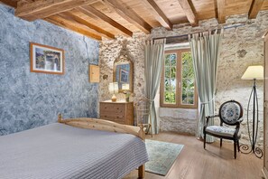 A rustic bedroom with a wooden bed, stone walls, a dresser, a chair, and a window with curtains.