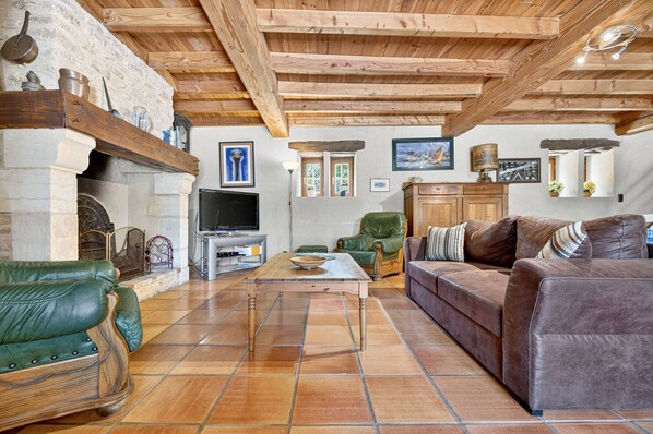 A rustic living room with exposed wooden beams, a stone fireplace, leather sofas, and a tiled floor.