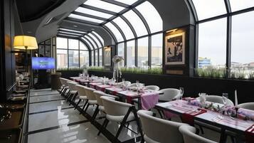 A restaurant with a curved ceiling and large windows offering a view of the city. Tables are set with white chairs and pink tablecloths.