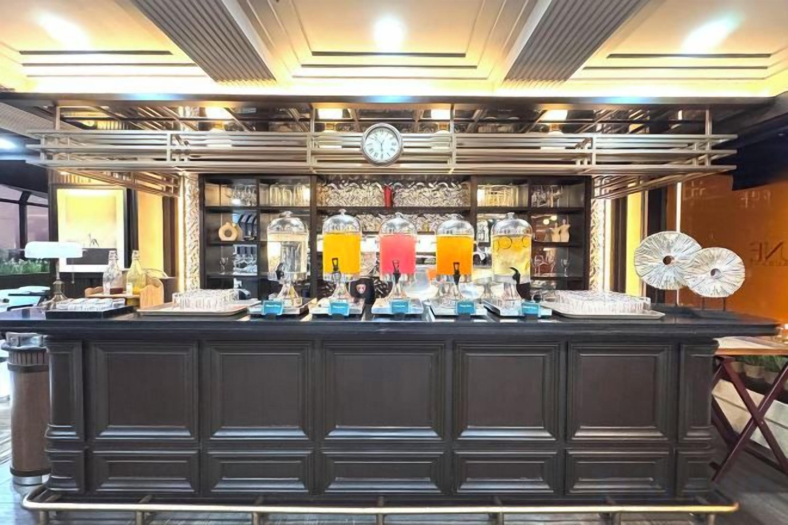 Zona de bar con mostrador oscuro, varios dispensadores de bebidas y un reloj en la pared.