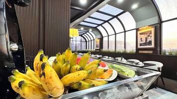 A buffet area with a variety of fruits including bananas and watermelon, and a view of the city through a glass ceiling.